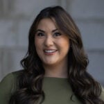 Sydnee Olsen, wearing a green top and with long wavy brown hair, smiles in front of a blurred gray background on the Restoration Webmasters site.